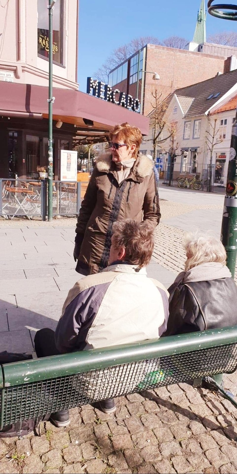 Daglig leder på Sammensenteret i Kristiansand, Hervor Julsrud, står foran to mennesker på en benk i en tom bygate.